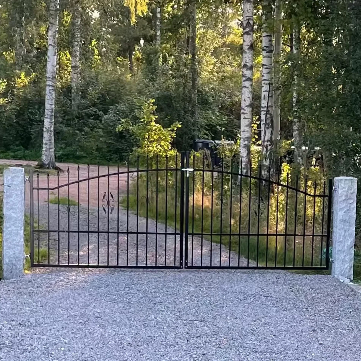 Svart dubbelgrind i smide mellan stenstolpar vid en grusinfart